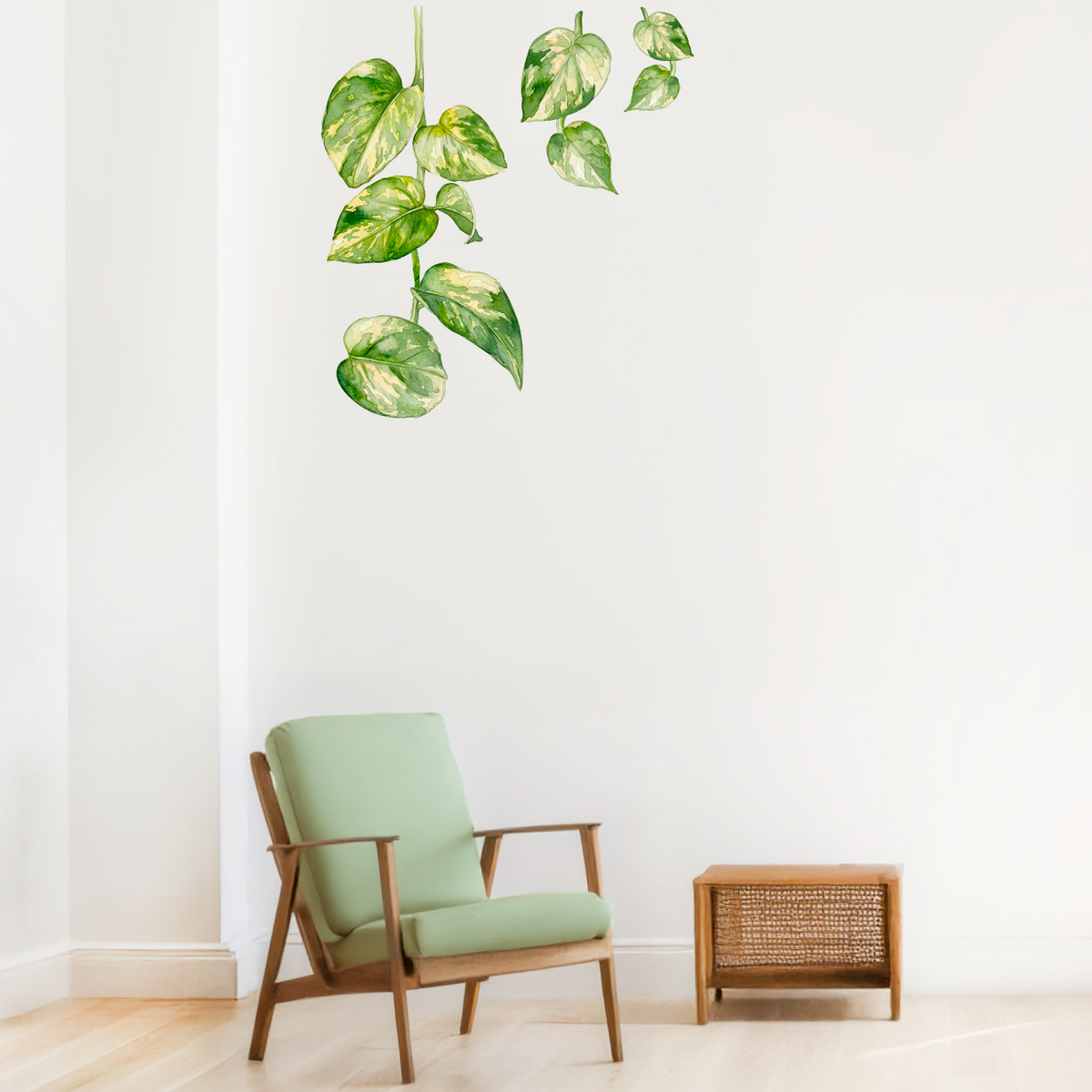A wall decal of green variegated pothos plant leaves applied to a white wall in a modern room setting with a light green armchair and a small wooden side table.